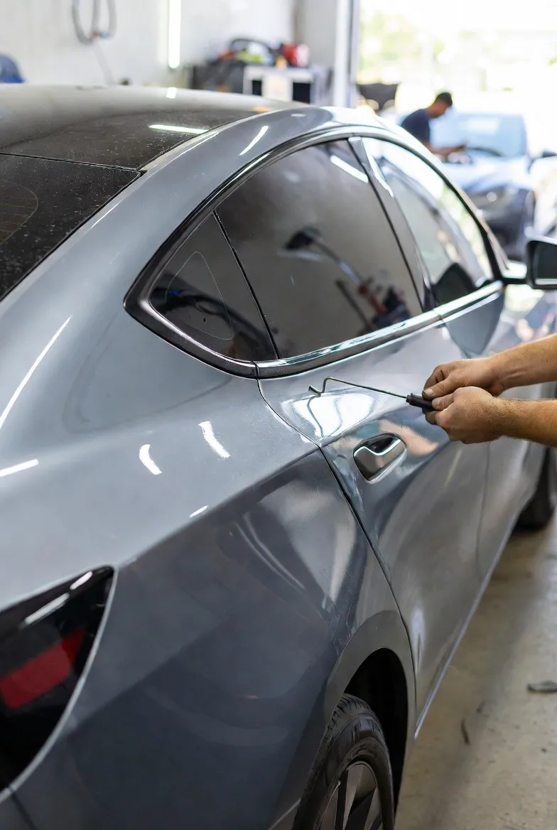 Mechanic using paintless dent repair tools to fix a dent on a gray car door in a workshop